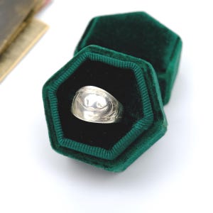 Peut inclure: Une bague en argent avec un motif gravé est présentée dans un écrin en velours vert foncé. La bague a un dessus arrondi et un motif détaillé autour de la bande. L'écrin est ouvert, révélant la bague sur un intérieur noir.
