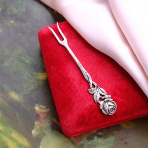 Puede incluir: Un tenedor de servir plateado con un diseño de rosa y hojas. El tenedor tiene dos púas y un motivo floral detallado en la base. Descansa sobre una superficie de terciopelo rojo, con una tela rosa suave en el fondo.