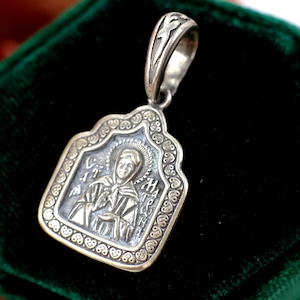 May include: A silver pendant with a religious icon design. The pendant features a detailed image of a saint with a halo, framed by an ornate border. The bail is also silver and has a decorative pattern.