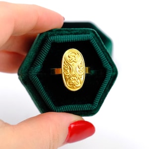 May include: A gold ring with an oval, ornate design is displayed in a dark green velvet ring box. The ring's central design features swirling patterns. The ring is held in a hexagonal box, creating a striking contrast.