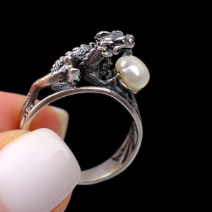 May include: A silver ring featuring a detailed, textured dragon figure wrapping around the band. The dragon's head is positioned to hold a single, smooth, white, oval-shaped pearl in its mouth. The metal has a dark, oxidized finish.