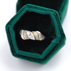 Può includere: Un anello color argento con una pietra centrale trasparente taglio marquise, affiancata da due pietre trasparenti più piccole su ciascun lato. La fascia ha una finitura liscia e lucida, presentata all'interno di un cofanetto esagonale in velluto verde.