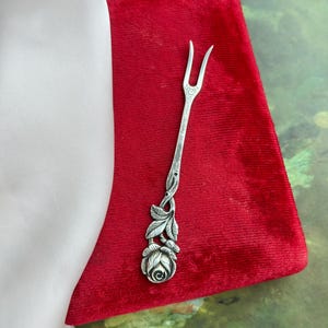 May include: A silver-toned serving fork with a floral rose design. The fork has two tines and a stem that is decorated with leaves and a rose at the base. The fork is resting on a red velvet surface.