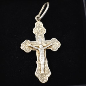 Peut inclure: Pendentif en argent en forme de croix, orné d'un motif élaboré avec une représentation de Jésus-Christ. La croix présente des motifs décoratifs et une boucle pour la suspension. Le fond est de couleur sombre.