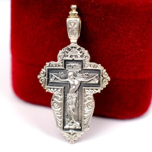 Puede incluir: Colgante de plata con forma de cruz, con un diseño ornamentado y una representación de Jesucristo. La cruz tiene detalles intrincados y una argolla para colgar. El fondo es de color rojo oscuro.