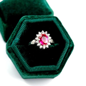 May include: A silver ring featuring a central oval, ruby-red gemstone encircled by small, clear stones. The ring is presented in a dark green velvet hexagonal ring box.