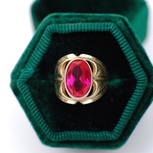 May include: A gold ring with a large, oval-cut, bright red gemstone. The ring is set in a gold band and is displayed in a dark green velvet ring box. The gemstone is the focal point of the ring.