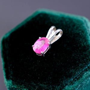 May include: A silver pendant with a bright pink, oval-shaped gemstone. The gemstone is set in a silver frame with a loop for a chain. The pendant is displayed on a dark green velvet surface.