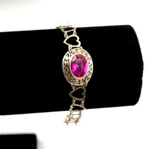 May include: Silver bracelet featuring an oval, magenta gemstone set in an ornate, filigree design. The bracelet's chain incorporates heart-shaped links, creating a romantic and elegant aesthetic. The bracelet is displayed against a black background.