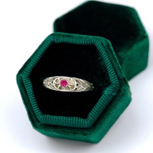 May include: A silver ring featuring a small, round, vibrant pink gemstone set in the center of an oval-shaped, textured metal frame. The ring is displayed inside a hexagonal, dark green velvet box with a black interior lining.