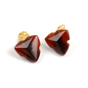 May include: A pair of clip-on earrings with a geometric design. The earrings are a deep reddish-brown colour with a faceted surface. The clip-on mechanism is gold-coloured and features a decorative design. The earrings are set against a white background.