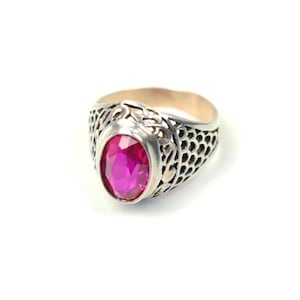 May include: A silver-toned ring featuring an oval-shaped, faceted hot pink gemstone set in a metal band with an intricate, openwork honeycomb or scale-like pattern that extends around the sides of the ring, creating a textured appearance.