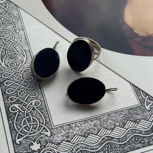 May include: A set of three silver rings with black oval stones. The rings are on a white surface with a black and white patterned border.
