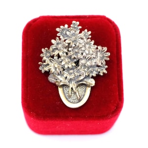 Puede incluir: Un broche de metal plateado con forma de ramo de margaritas dispuesto en un pequeño jarrón ovalado. El diseño floral presenta múltiples cabezas de flores texturizadas con pétalos y tallos detallados, acabados con una pátina oscura oxidada.
