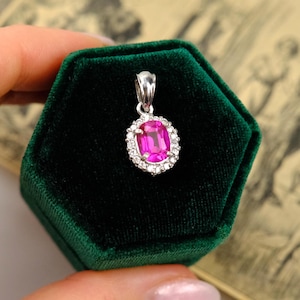 Puede incluir: Un colgante de plata con una vibrante gema rosa en el centro, rodeada por un halo de pequeñas piedras blancas brillantes. El colgante se exhibe sobre una superficie de terciopelo verde oscuro, creando un contraste llamativo. La gema es de forma ovalada.