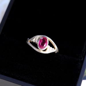 May include: A silver ring featuring a central oval-cut vibrant pink gemstone. The band has a split design that curves around the stone, creating an eye-like shape. The metal has a polished finish, and the setting holds the stone securely in place.