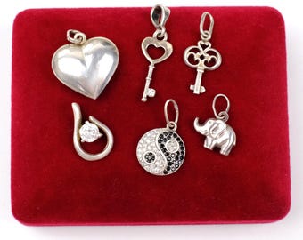 May include: Collection of silver pendants displayed on a red velvet surface. The pendants include a heart, keys, a teardrop with a clear stone, a yin-yang design, and an elephant. Each pendant has a loop for a chain.