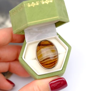 May include: An oval ring featuring a polished tiger's eye gemstone with brown and golden bands, set in a simple silver-toned metal band. The ring is displayed inside an open, hexagonal, light green velvet jewelry box with white lining.