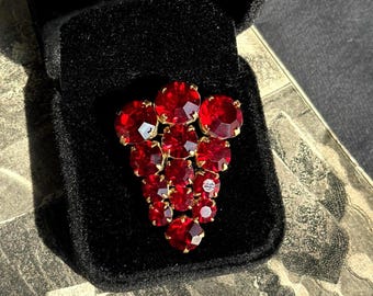 May include: A vintage brooch featuring a cluster of red, faceted gemstones set in a gold-tone metal. The brooch is shaped like a bunch of grapes and is displayed in a black velvet box.