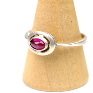 May include: A silver-toned ring featuring a smooth, oval-shaped, deep magenta cabochon gemstone set within a polished metal bezel. The band is slender and tapers slightly as it connects to the central oval setting, creating a simple, clean look.