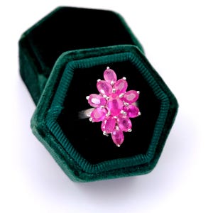 May include: A silver ring featuring a cluster of eleven oval-cut vibrant pink gemstones arranged in a floral-like pattern. The ring is displayed inside a dark green hexagonal velvet box with a soft, textured interior lining and a matching lid.