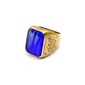 Puede incluir: Un anillo de tono dorado con una gran piedra de vidrio azul profundo de forma rectangular engastada en el centro. La banda de metal es ancha y muestra un patrón floral grabado en el lateral, con líneas decorativas grabadas alrededor.