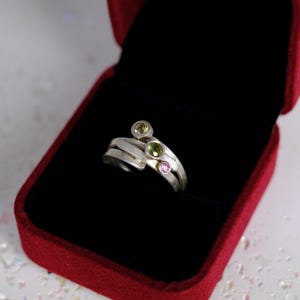 May include: A silver ring with three gemstones: a light green, a dark green, and a pink stone. The ring is displayed in a red velvet ring box with a black interior. The ring has a unique, layered design.