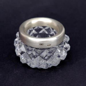 Puede incluir: Un pequeño salero de cristal transparente y forma redonda con un patrón de facetas talladas en diamante en el exterior. El borde superior tiene una banda de plata de acabado mate liso con el texto estampado M1.9 y un pequeño sello adicional.