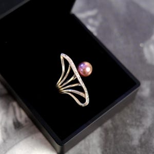 May include: A silver ring featuring a fan-shaped design with small embedded stones along the outer edge and a single round, iridescent purple pearl positioned at the top of the structure, resting inside a black jewelry box.
