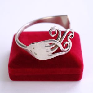 May include: A silver-toned bracelet crafted from a fork, featuring the fork's tines and a decorative swirl design. The bracelet is displayed on a red velvet surface, showcasing its unique, handcrafted style. The bracelet's design is a creative repurposing of a common utensil.