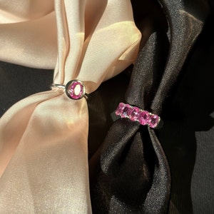 May include: Two silver rings with pink gemstones. One ring has a single large oval gemstone, the other has a row of smaller oval gemstones.