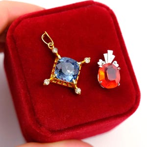 May include: Two gemstone pendants. One pendant features a large, square, light blue gemstone set in a gold-coloured frame with small, clear accent stones. The other pendant has a red-orange gemstone with a silver-coloured setting.