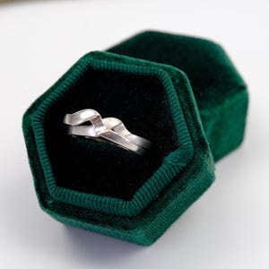 Puede incluir: Un anillo de plata con un diseño único e entrelazado descansa dentro de un joyero de terciopelo verde oscuro. El joyero está abierto, revelando el anillo contra un interior negro. La caja tiene forma hexagonal.