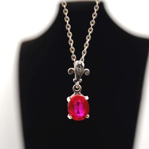 May include: Silver-toned pendant necklace featuring a vibrant red oval gemstone. The pendant has an ornate design with a fleur-de-lis element and a delicate chain. The necklace is displayed against a black background.