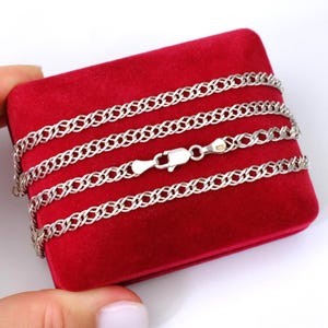 May include: A silver-toned chain necklace featuring a repeating interlocking link pattern. The chain is arranged in multiple loops and includes a secure lobster claw clasp mechanism, showcasing a polished metallic finish throughout the design.