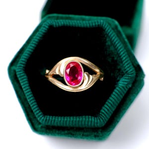 May include: A gold ring with a large, oval-cut ruby gemstone set in a decorative setting. The ring is displayed in a dark green velvet ring box, creating a striking contrast. The ring's design features elegant, flowing lines.
