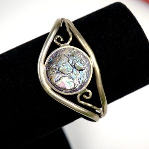 May include: Silver cuff bracelet with a round abalone shell centrepiece. The iridescent shell is set within a decorative frame with swirling accents. The bracelet is displayed on a black surface.