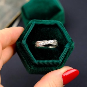 Puede incluir: Un anillo de plata con cinco pequeñas piedras claras engastadas en un diseño ondulado. El anillo se muestra en una caja hexagonal de terciopelo verde oscuro. Joyería.