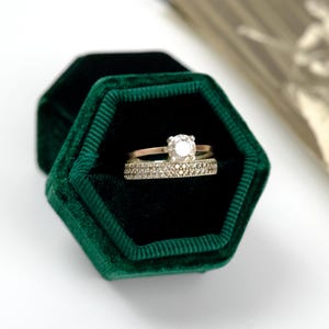 May include: A gold engagement ring with a large, clear center stone and a diamond band sits inside a dark green velvet hexagonal ring box. A second ring with a diamond band is also in the box. The box is open, revealing the black interior.