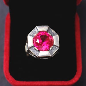 Puede incluir: Un anillo de plata con una gran gema rosa brillante facetada. El anillo tiene un diseño geométrico alrededor de la piedra, sobre un fondo negro dentro de una caja roja. Una pieza llamativa.