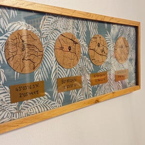 May include: A rectangular wooden framed wall art piece. The artwork features four circular cork maps set against a teal background with a white leaf pattern. Each map has coordinates on a small brass-coloured plaque.