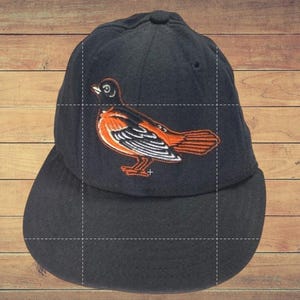 May include: Black baseball cap featuring an embroidered orange and black bird design. The bird is detailed with white accents. The cap has a curved brim and a rounded crown. The background is a wooden surface.