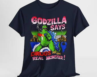 Könnte beinhalten: Ein marineblaues Kurzarm-T-Shirt mit einer Grafik eines grünen Monsters, das eine große rot-blaue Pille hält. Der Text lautet GODZILLA SAYS DRUGS ARE THE REAL MONSTER! Der Hintergrund zeigt eine Skyline, Blitze und einen grünen Mond.