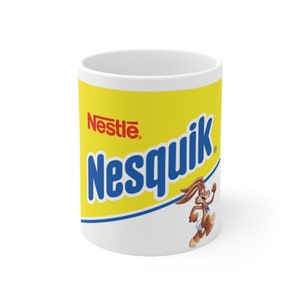 Puede incluir: Taza de cerámica blanca con el logotipo de Nesquik en azul y el logotipo de Nestlé en rojo sobre un fondo amarillo. También presenta una mascota de conejo de dibujos animados.