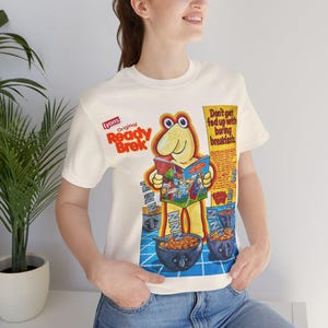 Puede incluir: Camiseta color crema con un diseño vintage de cereales "Ready Brek". El diseño incluye un personaje de dibujos animados leyendo un cómic, cuencos de cereales y el texto "Don't get fed up with boring breakfasts."