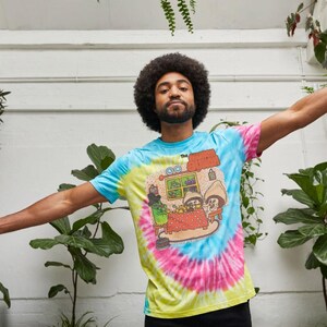 May include: A tie-dye t-shirt with a colorful design featuring a cartoon scene with mushroom characters. The shirt has a blue, yellow, and pink spiral pattern. The text "The Shroom" is visible on the shirt.