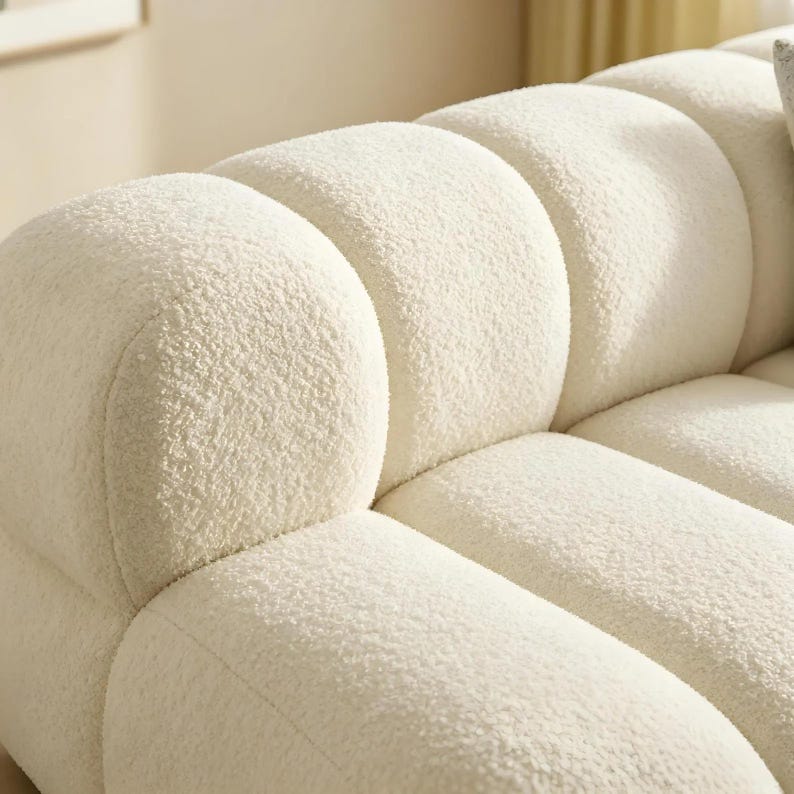 May include: Close-up of a cream-colored, textured sofa. The sofa has a rounded, cloud-like design with a soft, plush appearance. The fabric has a textured, boucl&eacute; finish, creating a cozy and inviting aesthetic.