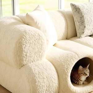 May include: Cream-colored, textured sofa with a built-in cat bed. The sofa has a rounded design and includes two throw pillows. A tabby cat is inside the cat bed. The sofa is near a window.