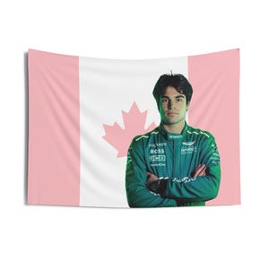 May include: A Canadian flag with a pink background and a white maple leaf. A man wearing a green racing suit with the text "Aramco", "Bachant", "BOSS", "JCB", and "InfiniteOne" is standing in front of the flag.