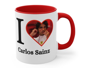 May include: White ceramic mug with a red rim and handle. The mug has a black "I" and a red heart with a picture of Carlos Sainz inside. The text "Carlos Sainz" is printed below the heart.